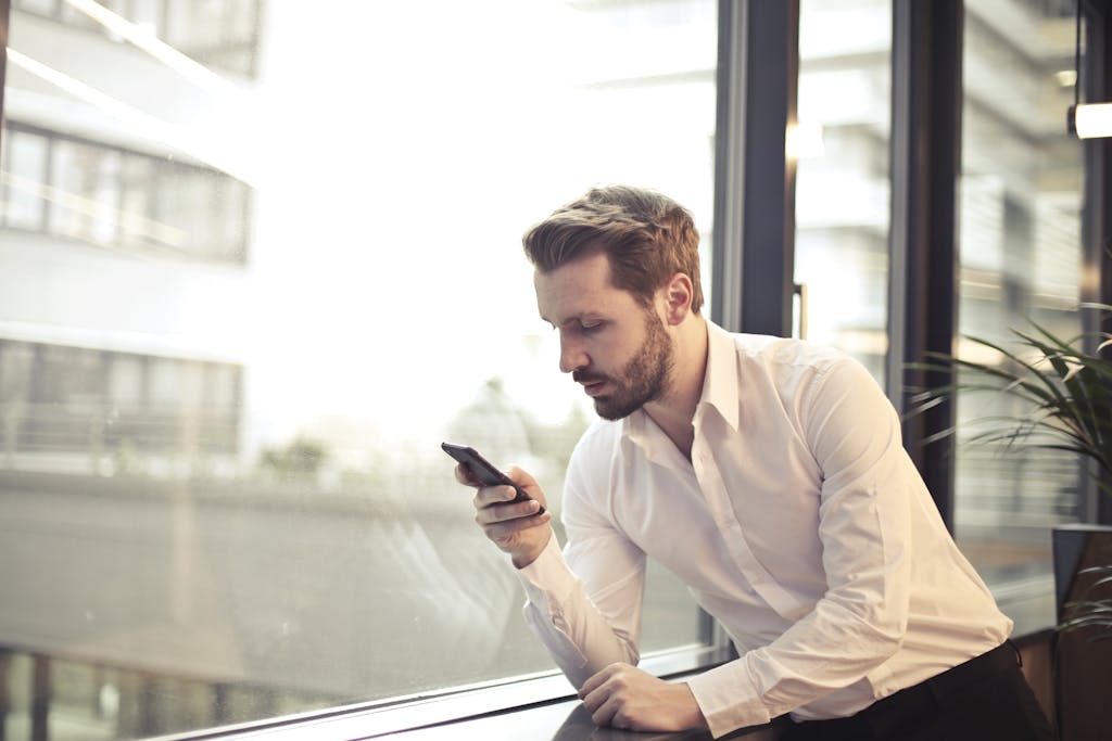 A businessman in a white shirt texts on his smartphone by a large glass window inside an office.
